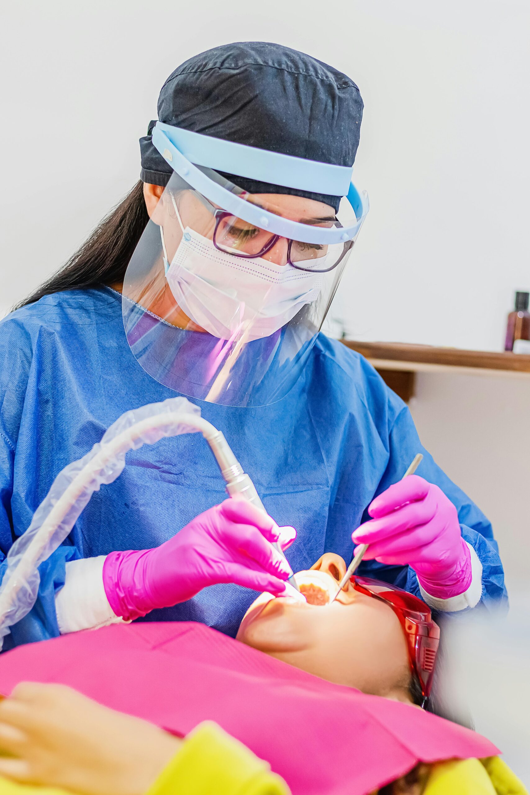 A female dentist wearing protective gear performs a dental procedure on a patient in a clinic setting.